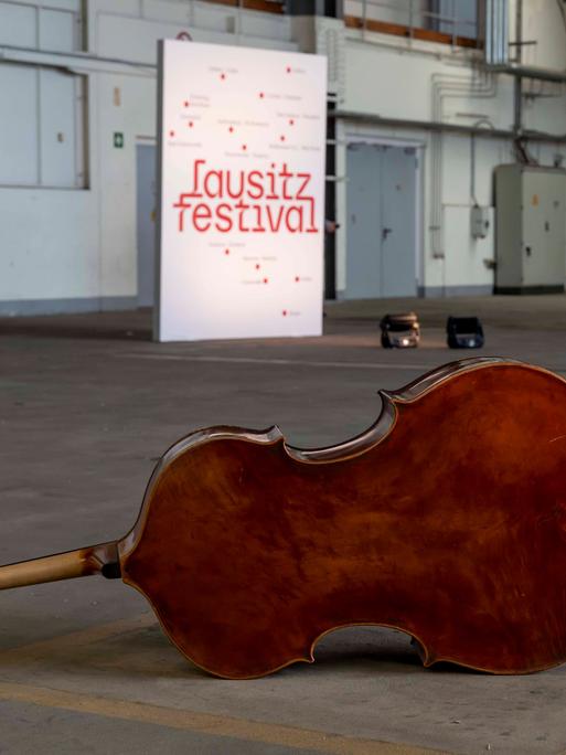 In einem fast leeren Hanger sind im Hintergrund ein weißes stehendes Banner mit der roten Aufschrift "lausitz festival" und im Vorgergrund ein liegender Kontrabass zu sehen.