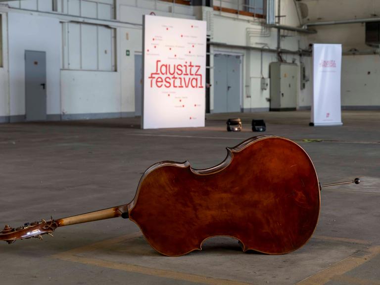 In einem fast leeren Hanger sind im Hintergrund ein weißes stehendes Banner mit der roten Aufschrift "lausitz festival" und im Vorgergrund ein liegender Kontrabass zu sehen.