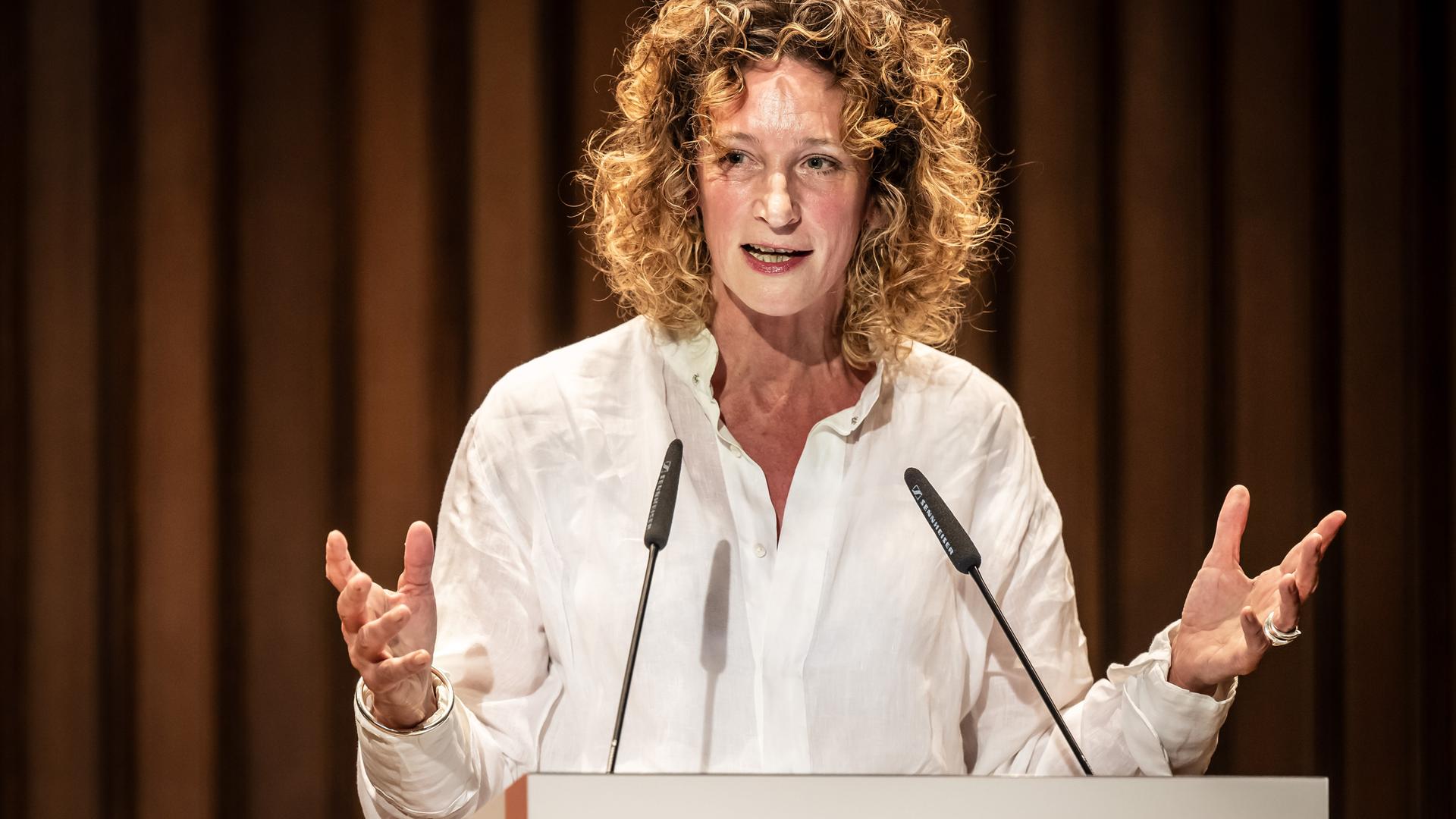 Wiebke Puls steht an einem Pult vor einem Theatervorhang und spricht ins Mikrofon. Wiebke Puls steht an einem Pult vor einem Theatervorhang und spricht ins Mikrofon.