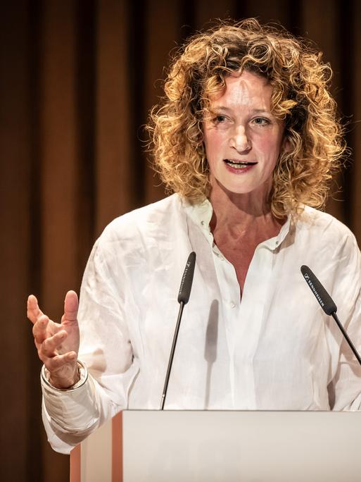 Wiebke Puls steht an einem Pult vor einem Theatervorhang und spricht ins Mikrofon.