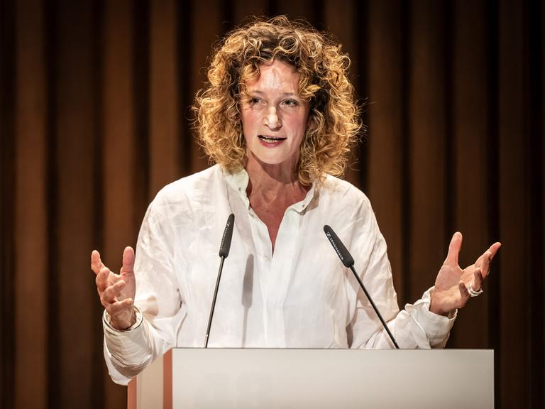 Wiebke Puls steht an einem Pult vor einem Theatervorhang und spricht ins Mikrofon.