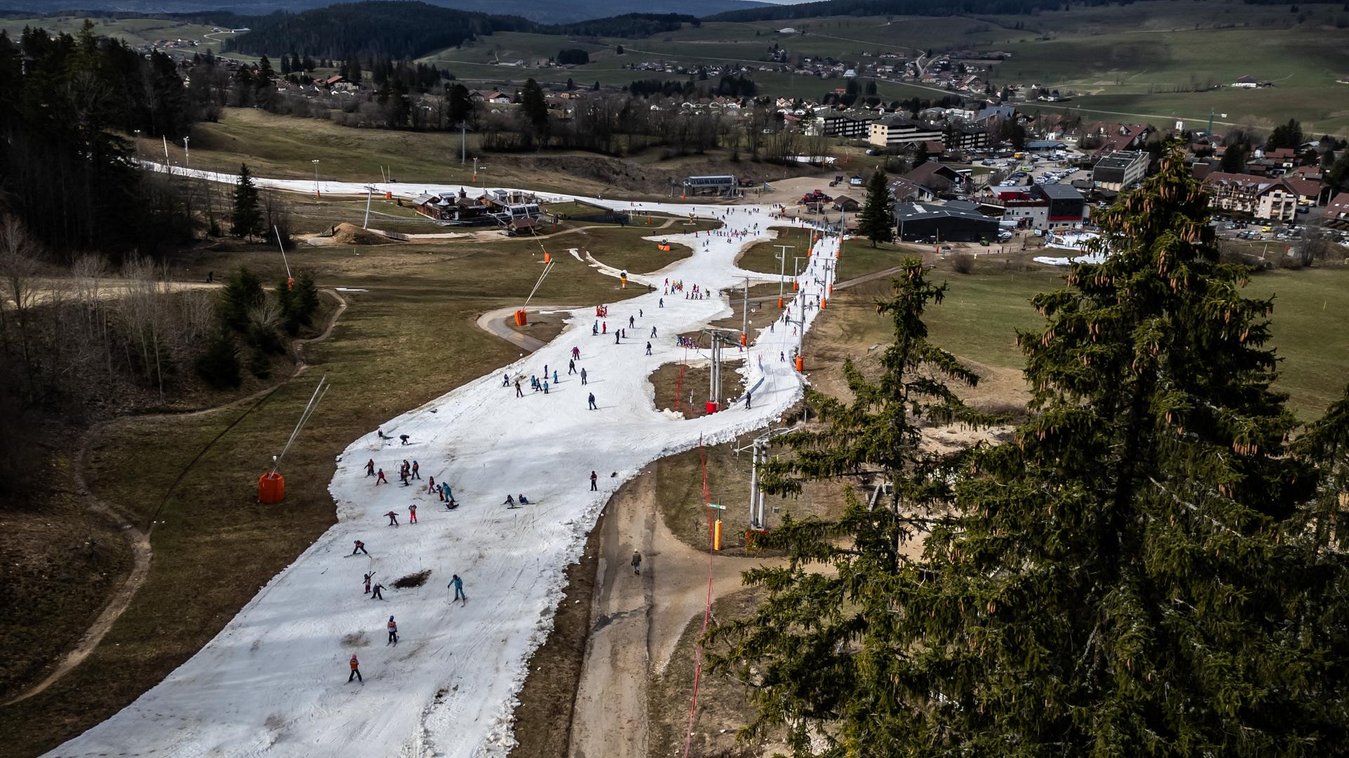 Eine Skipiste mit Skifahrern. Die Landschaft um die Piste ist klomplett schneefrei. Im Hintergrund ist eine Stadt zu sehen. 