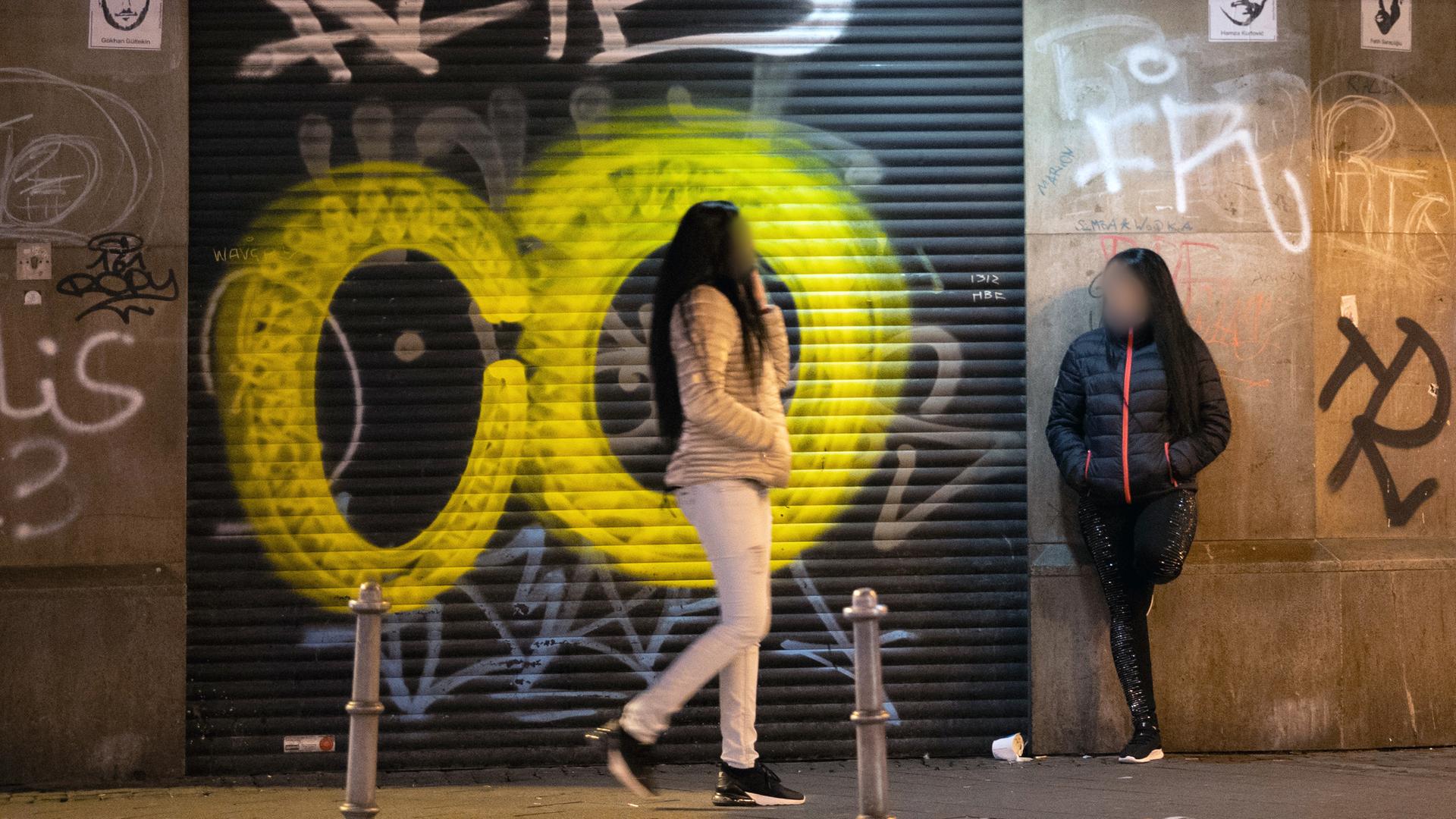 Zwei Prostituierte stehen im Frankfurter Bahnhofsviertel vor einem mit einem geschlossenen Torrolladen,  der ebenso wie die angrenzende Wand mit Graffiti-Tags besprüht ist.