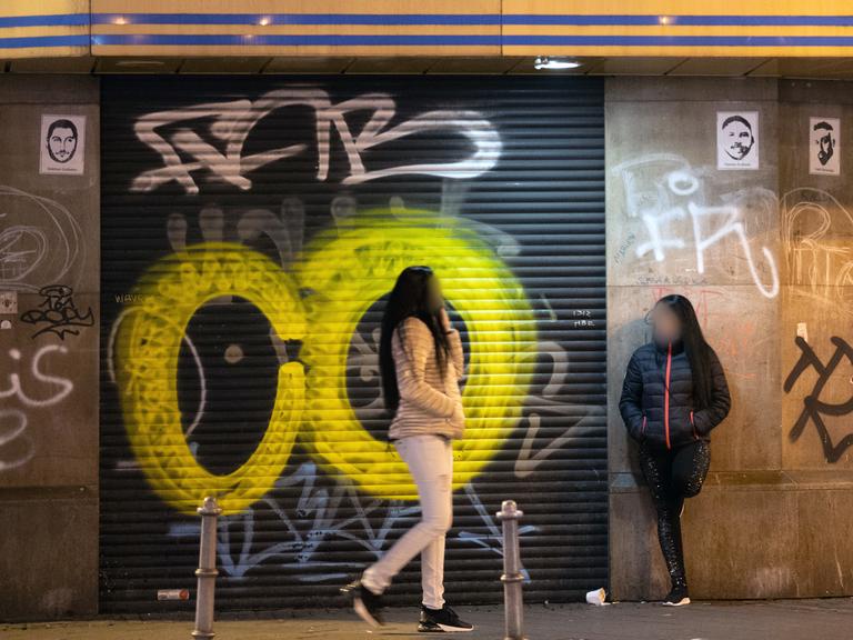 Zwei Prostituierte stehen im Frankfurter Bahnhofsviertel vor einem mit einem geschlossenen Torrolladen,  der ebenso wie die angrenzende Wand mit Graffiti-Tags besprüht ist.