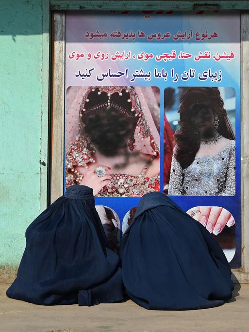 Zwei afghanische Frauen, in Burkas verhüllt, sitzen vor einem geschlossenen Schönheitssalon. Die Frauengesichter im Schaufenster sind mit schwarzer Farbe übersprüht worden. Jalalabad, 13.1. 2021.