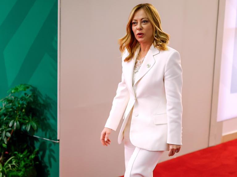 Italiens Regierungschefin Georgia Meloni läuft in einem weißen Hosenanzug einen Gang entlang. Italiens Regierungschefin Georgia Meloni läuft in einem weißen Hosenanzug einen Gang entlang.
