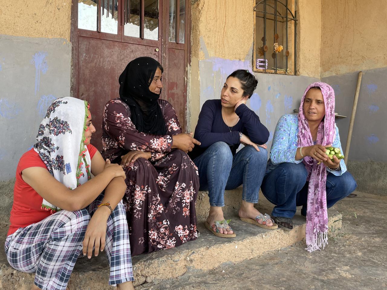 Auf der Treppe eines Hauses sitzen vier Frauen und unterhalten sich im Frauendorf Jinwar. Ganz rechts sitzt Zeyneb - bekleidet mit einer dunklen Jeans, einer hellblauen Bluse und einem rosa Kopftuch.