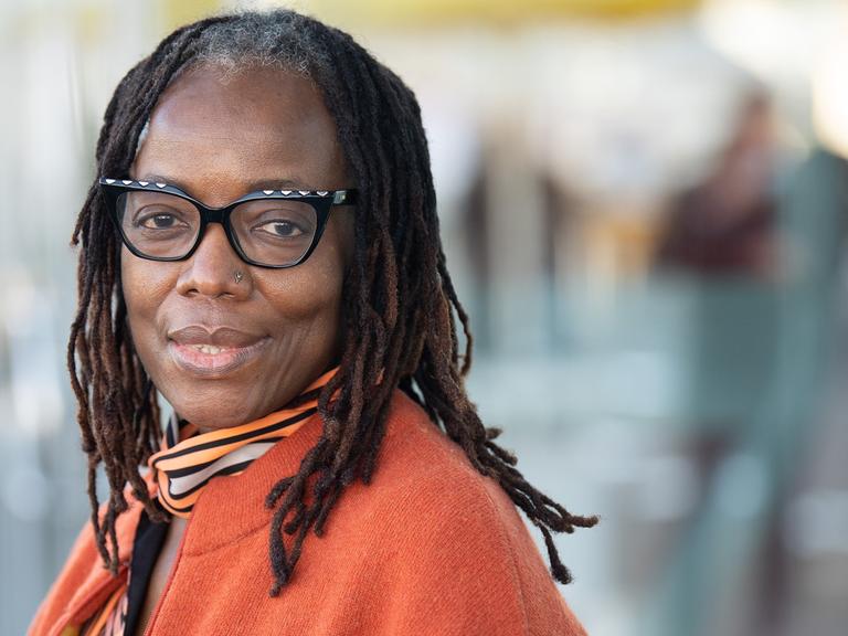 Porträtaufnahme der simbabwischen Autorin Tsitsi Dangarembga. Sie ist Trägerin des Friedenspreises des Deutschen Buchhandels 2021. Dangarembga trägt eine orangefarbene Jacke über einer orange-braun-weiß gemusterten Bluse. Ihre Haare trägt sie zu vielen Zöpfen geflochten.