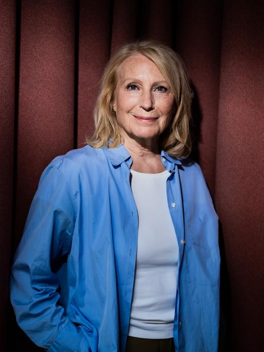 Maren Kroymann, Schauspielerin, steht während eines Fototermins in weißem T-Shirt und offenem blauen Hemd vor einem Vorhang und lächelt in die Kamera.