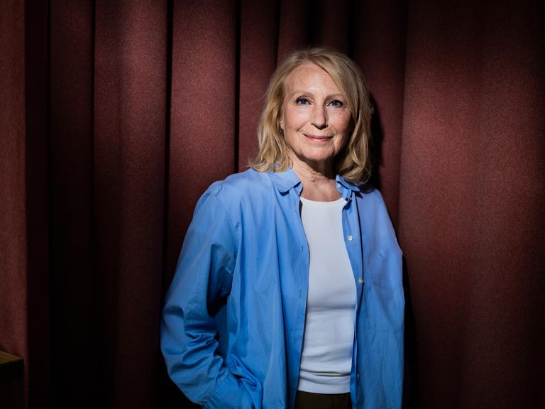 Maren Kroymann, Schauspielerin, steht während eines Fototermins in weißem T-Shirt und offenem blauen Hemd vor einem Vorhang und lächelt in die Kamera.