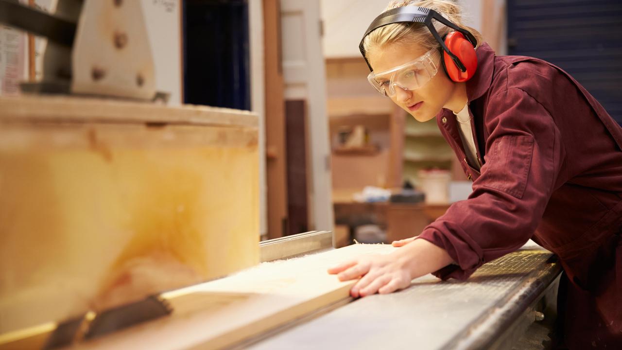 Eine junge Frau in Arbeitskleidung und mit Schutzbrille arbeitet an einer Kreissäge.