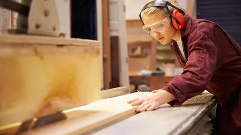 Eine junge Frau in Arbeitskleidung und mit Schutzbrille arbeitet an einer Kreissäge 