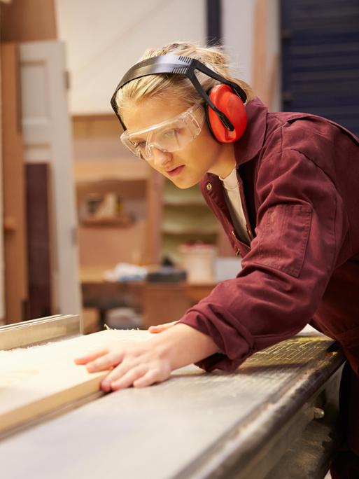 Eine junge Frau in Arbeitskleidung und mit Schutzbrille arbeitet an einer Kreissäge 