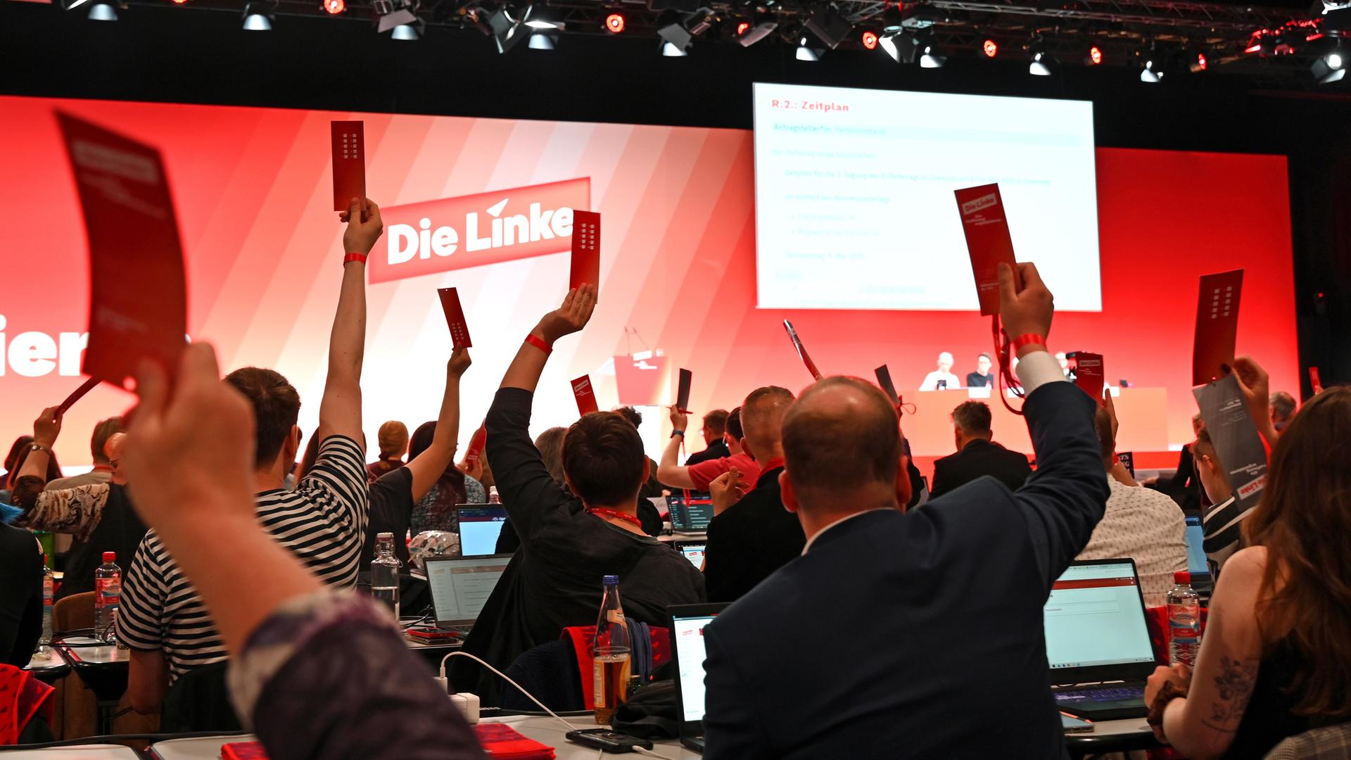 Blick in den Saal. Man sieht viele Delegierte von hinten in rötlichem Licht, die ihre Stimmkarten in die Höhe halten. I Hinteergrund eine rote Wand mit dem Parteilogo.