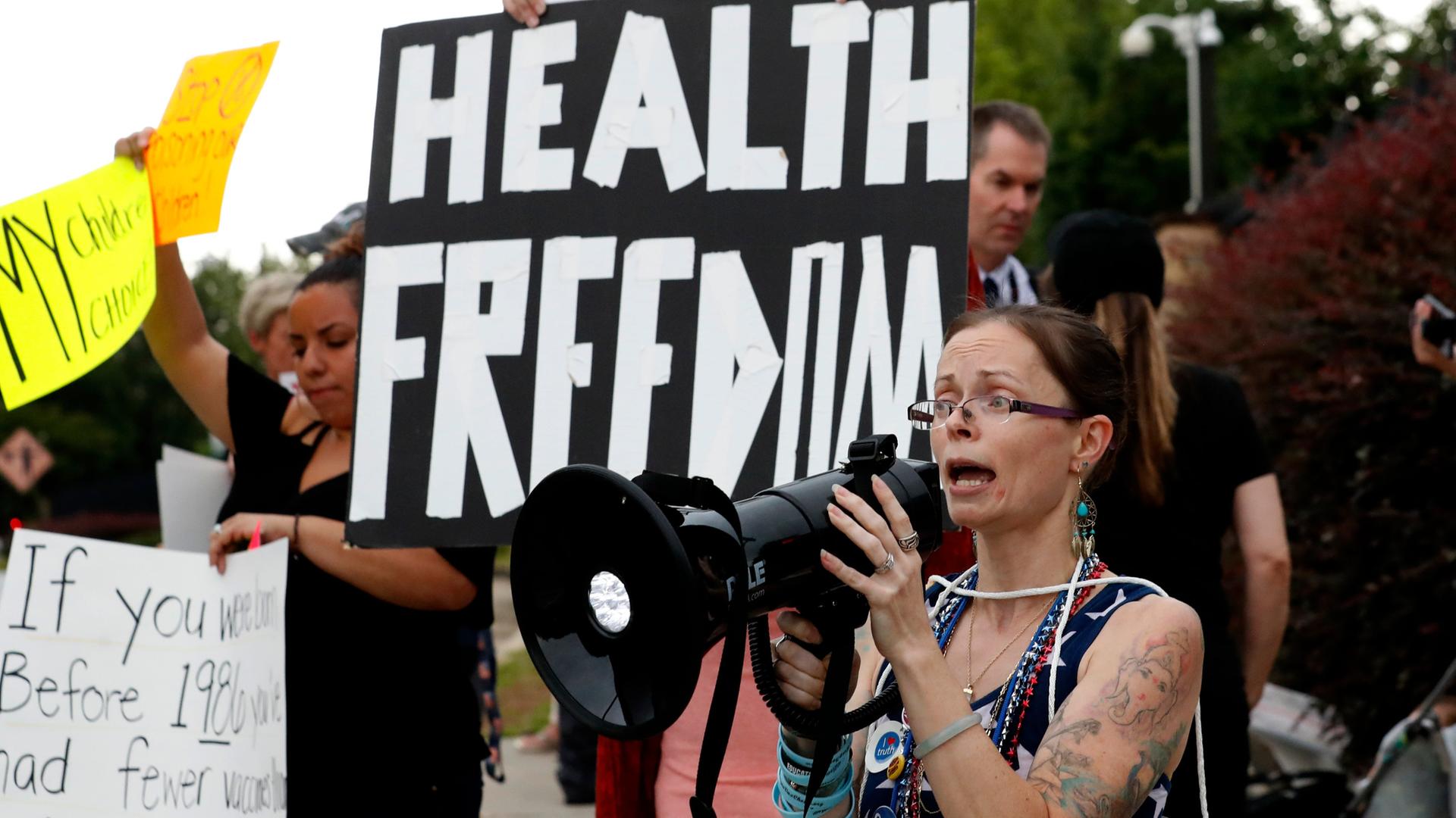 Eine Demonstrantin spricht in ein Megafon. Hinter ihr halten Leute ein Banner mit der Aufschrift "Health Freedom".