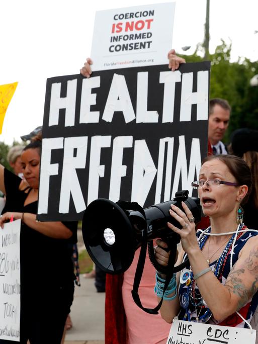 Eine Demonstrantin spricht in ein Megafon. Hinter ihr halten Leute ein Banner mit der Aufschrift "Health Freedom".