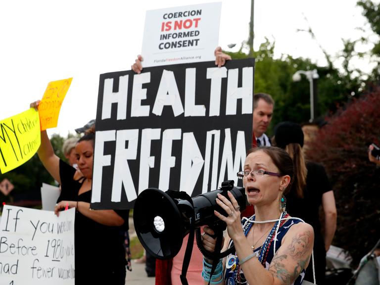 Eine Demonstrantin spricht in ein Megafon. Hinter ihr halten Leute ein Banner mit der Aufschrift "Health Freedom".