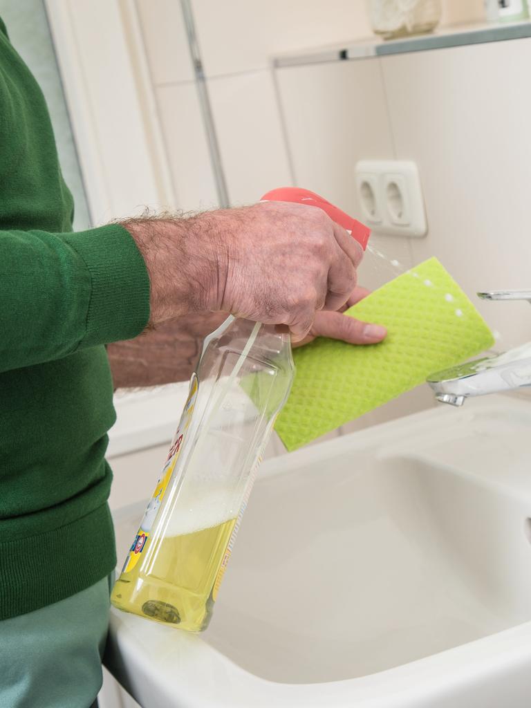 Ein Mann putzt ein Waschbecken mithilfe eines Lappens und Putzmittel.