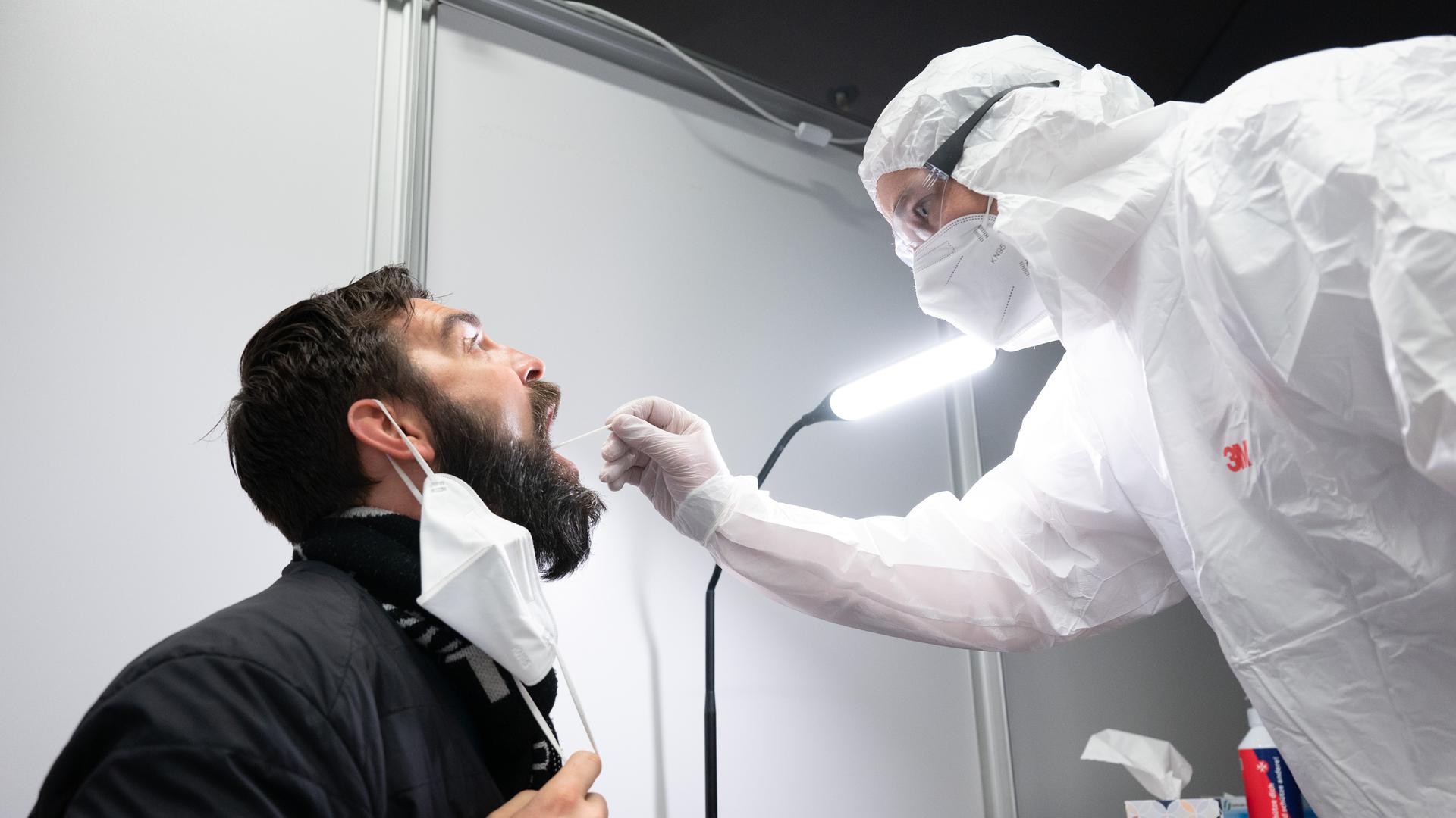 Ein Mann komplett in Sicherheitskleidung nimmt im aufgesperrten Mund eines anderen Mannes mit Bart die Testrobe. 