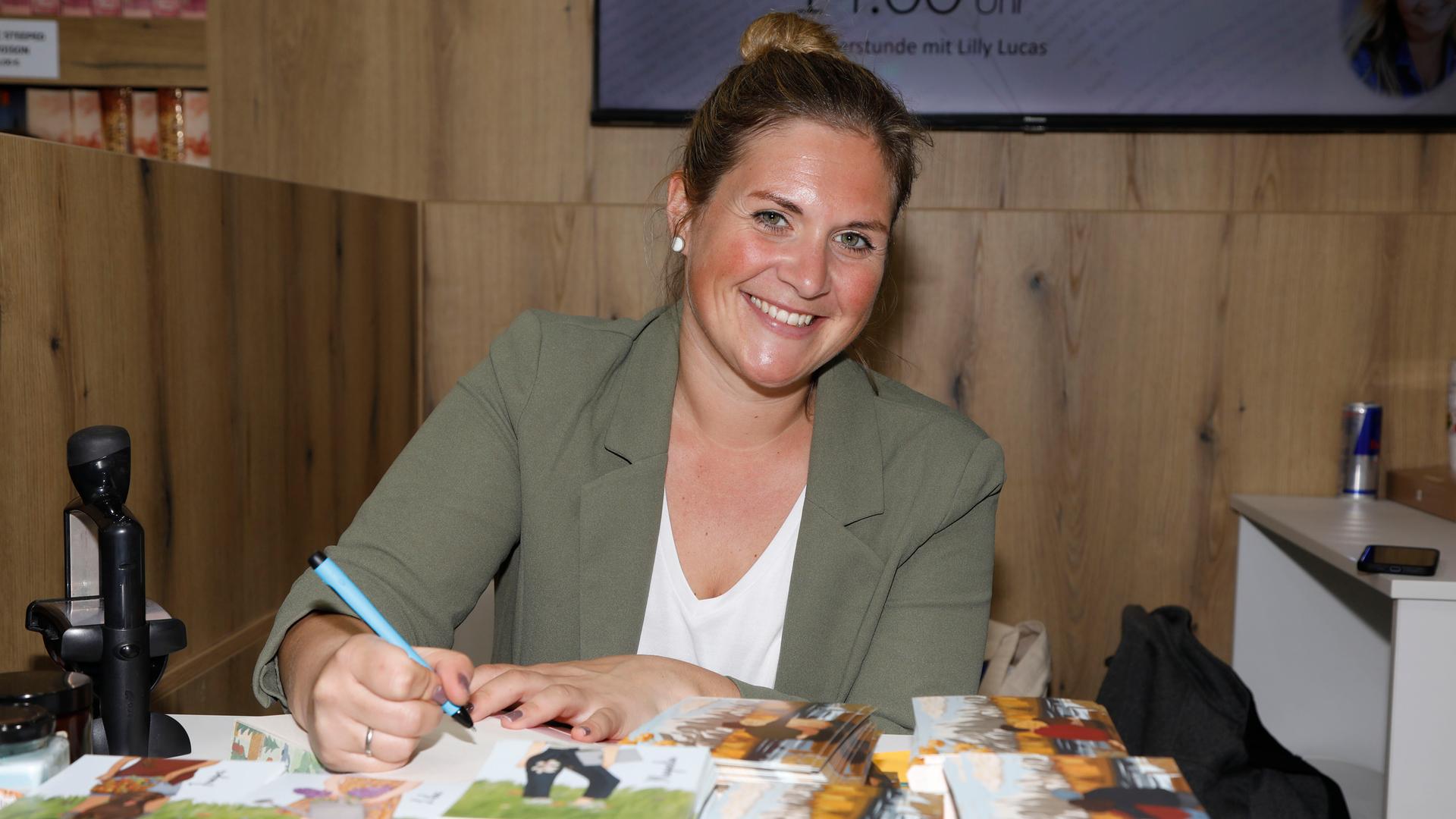 Die Autorin Lilly Lucas sitzt während der Leipziger Buchmesse an einem Stand und signiert eins ihrer Bücher, sie lächelt dabei in die Kamera.