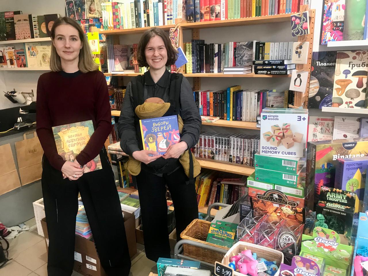 Zwei junge Frauen stehen inmitten von Büchern nebeneinander in einem Laden, in der Hand ebenfalls jeweils ein Buch.