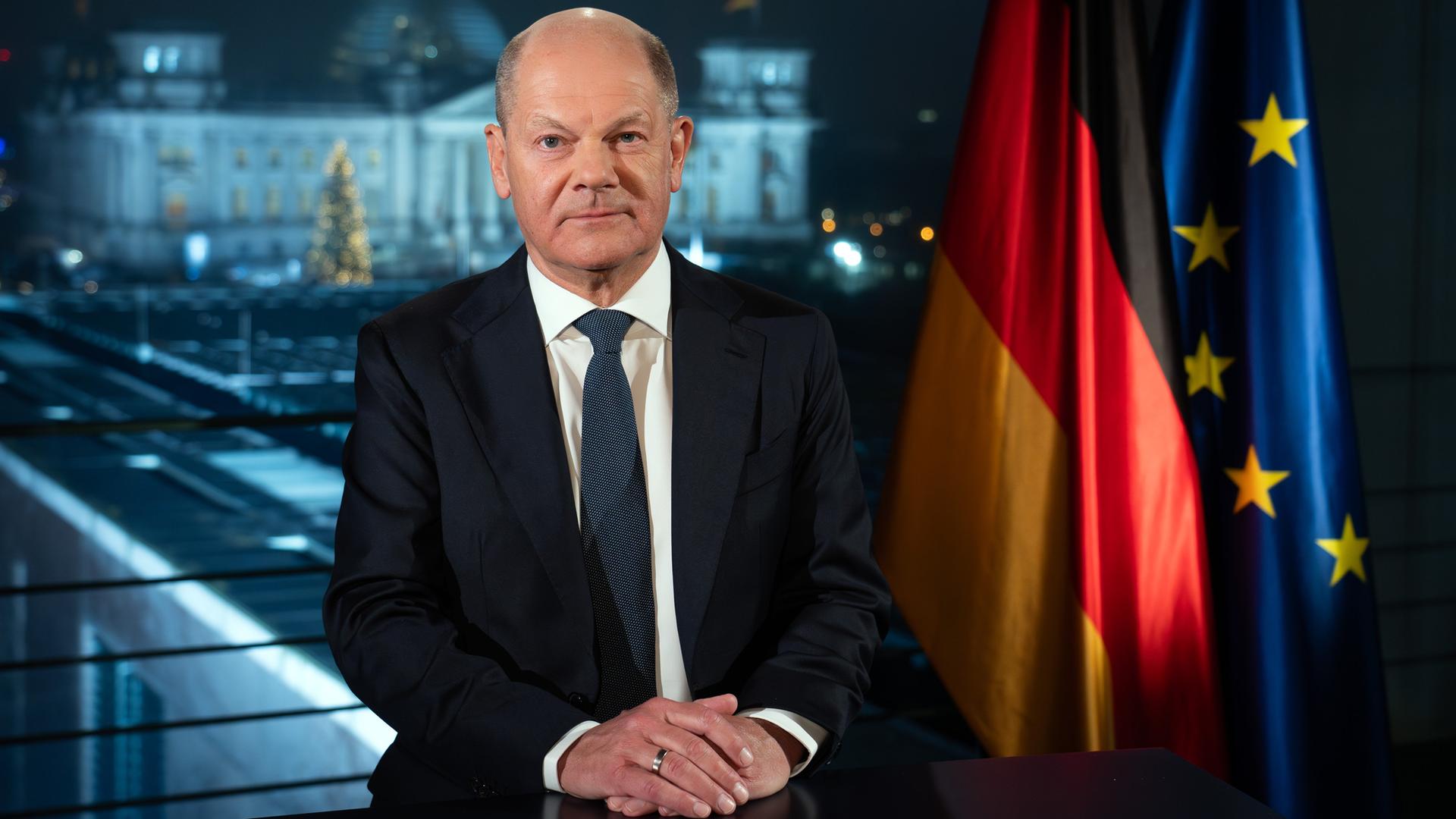 Olaf Scholz sitzt während eines Fototermins an einem Tisch im Kanzleramt vor der Kulisse des von Scheinwerfern angestrahlten Reichstages. Rechts hinter ihm sind auch eine Flagge Deutschlands und der Europäischen Union zu sehen.