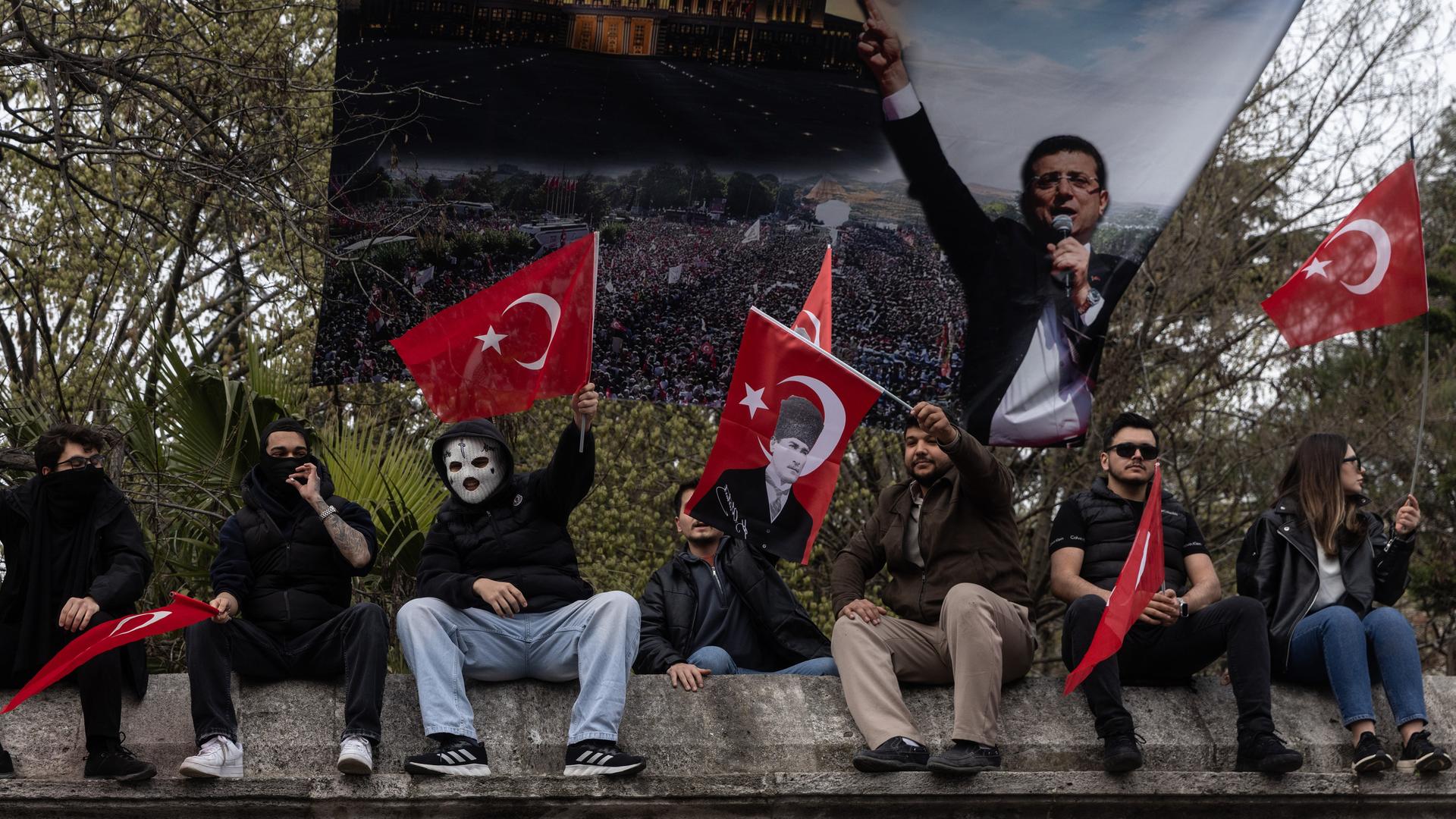 Eine Gruppe von Menschen sitzt auf einer Mauer, in deren Hintergrund ein Banner mit einem Foto von Ekrem Imamoglu zu sehen ist. Sie tragen teilweise Masken und sind nicht zu erkennen, einige wehen die türkische Flagge in der Luft.