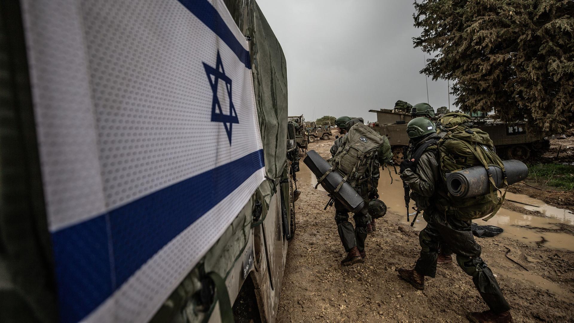 Israelische Soldaten in voller Ausrüstung gehen an einem Militärfahrzeug mit der israelischen Flagge vorüber.