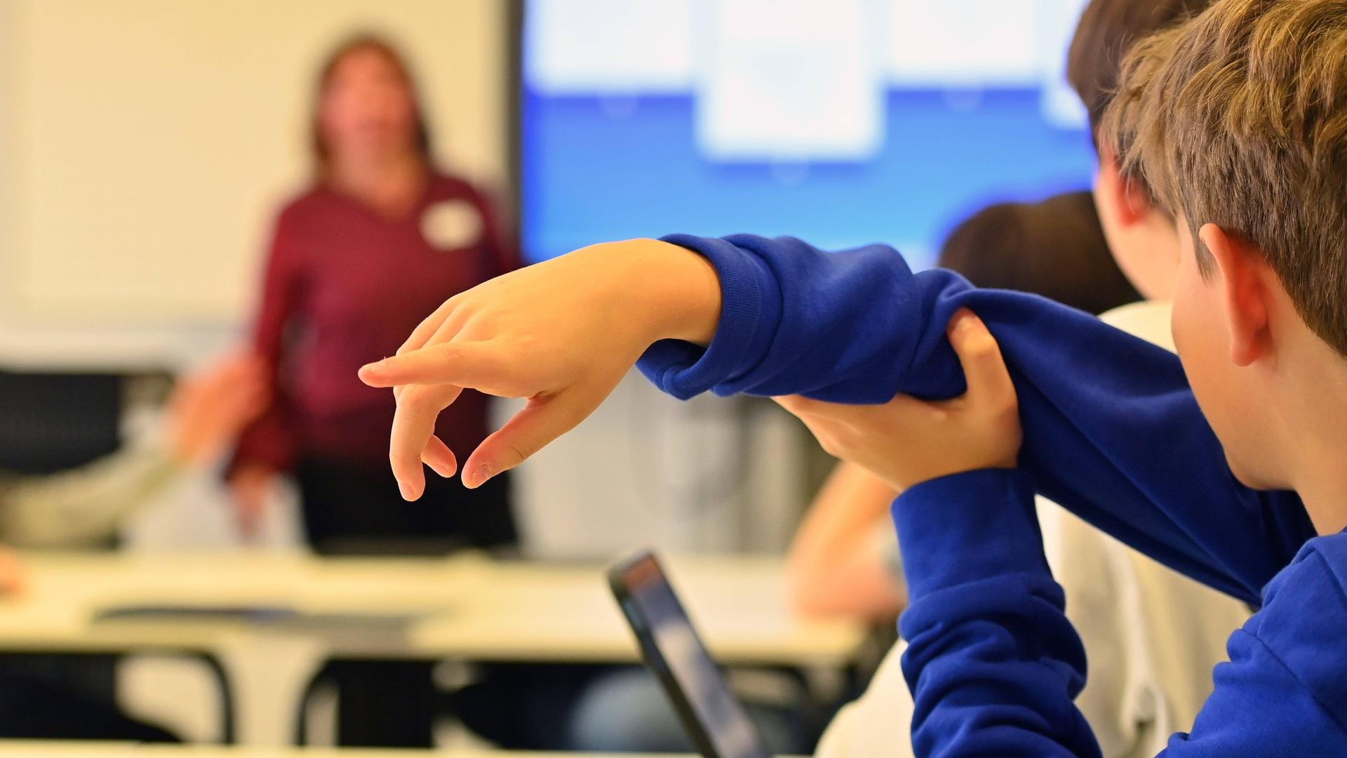 Ein Junge hebt in einem Klassenraum seine Hand. Im Hintergrund steht eine Lehrerin.