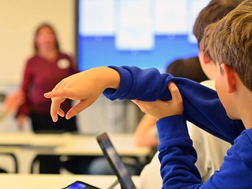 Ein Junge hebt in einem Klassenraum seine Hand. Im Hintergrund steht eine Lehrerin.