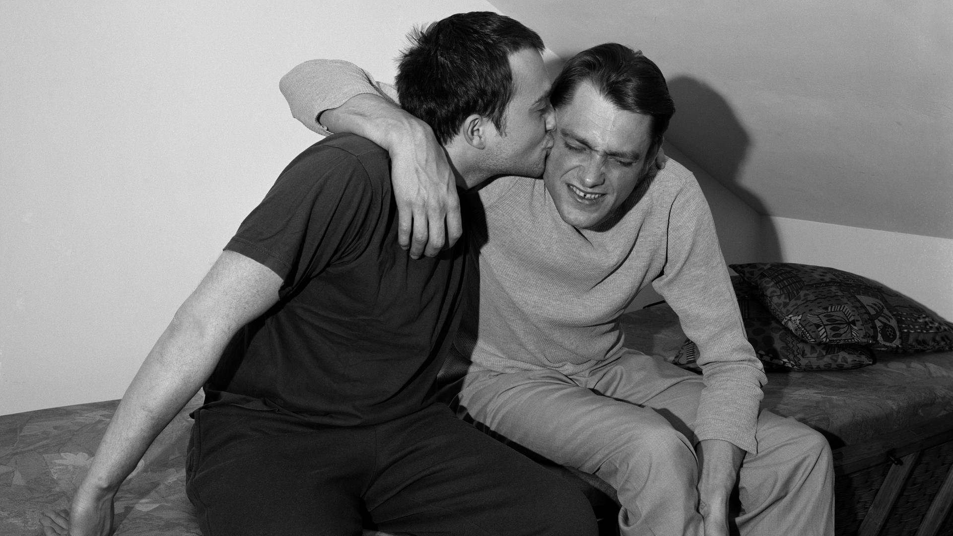 Schwarz-weiß Foto von einem Brüderpaar, das sich, auf einem Bett sitzend, in einer Dachkammer umarmt. Der eine Bruder küsst den anderen mit Begeisterung auf den Hals, der scheint nicht so angetan.