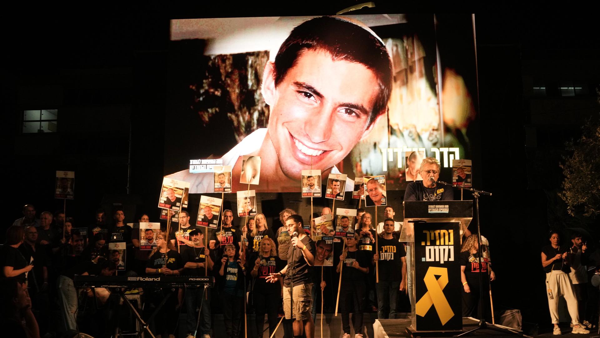 Es ist dunkel. Das lächelnde Gesicht Hard Goldins ist auf einer großen Leinwand zu sehen. Davor viele Demonstranten mit Plakaten, auf denen andere Geiseln abgebildet sind. Es ist dunkel. Das lächelnde Gesicht Hard Goldins ist auf einer großen Leinwand zu sehen. Davor viele Demonstranten mit Plakaten, auf denen andere Geiseln abgebildet sind.