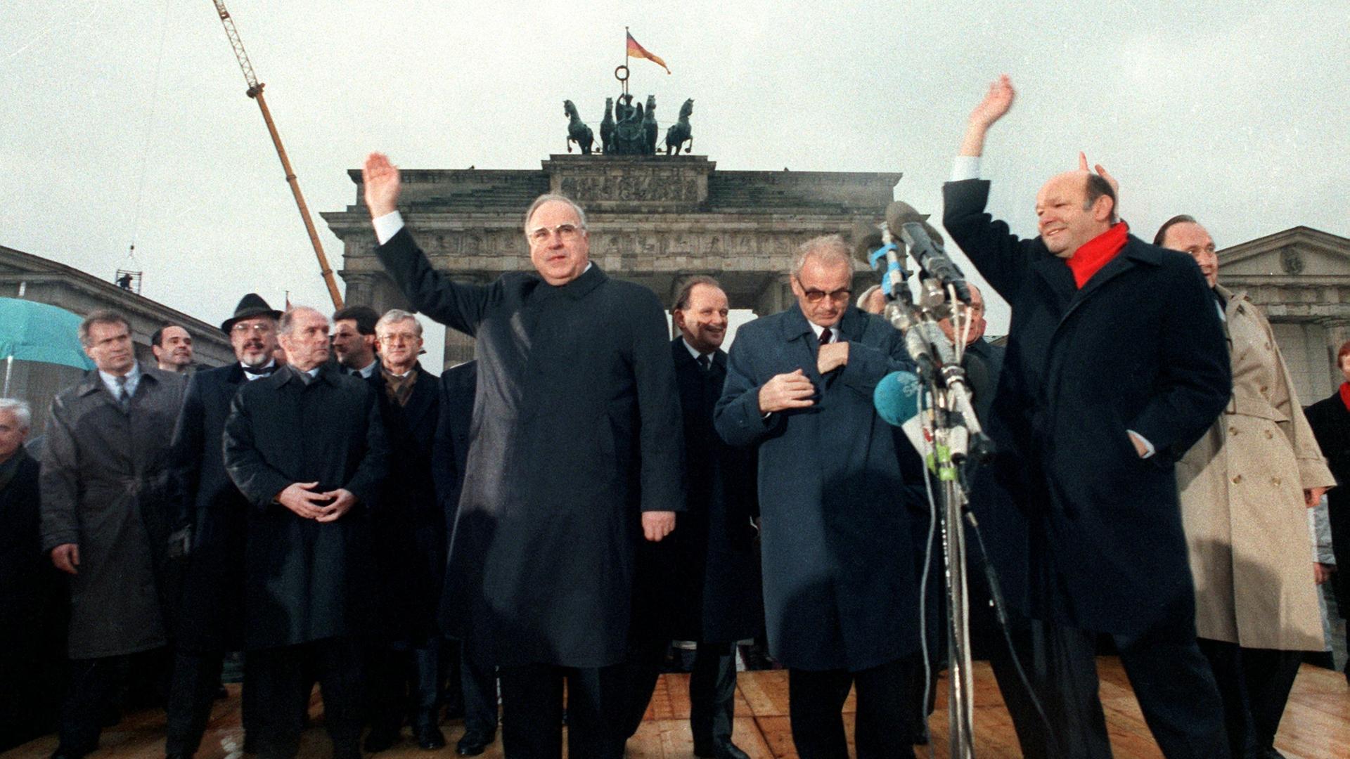 Bundeskanzler Helmut Kohl (l) und Berlins Regierender Bürgermeister Walter Momper (r) winken fröhlich der Menschenmenge zu. Zwischen ihnen der Ministerpräsident der DDR, Hans Modrow, im Hintergrund das Brandenburger Tor. 28 Jahre nach dem Bau der Mauer ist das Symbol der deutschen Teilung wieder zugänglich. Rechts und links neben dem Brandenburger Tor wurden Grenzübergänge geschaffen, die am 22. Dezember 1989 feierlich eröffnet wurden. [dpabilderarchiv]