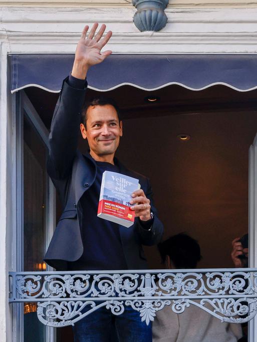Autor Jean-Baptiste Andrea winkt nach dem Gewinn des Prix Goncourt im November 2023 mit seinem Roman "Veiller sur elle" in der Hand von einem Balkon aus. 