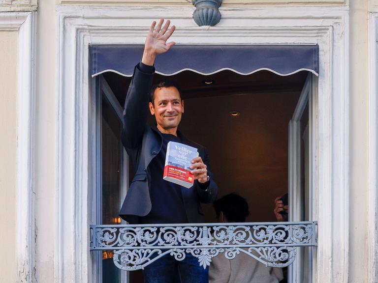 Autor Jean-Baptiste Andrea winkt nach dem Gewinn des Prix Goncourt im November 2023 mit seinem Roman "Veiller sur elle" in der Hand von einem Balkon aus. 