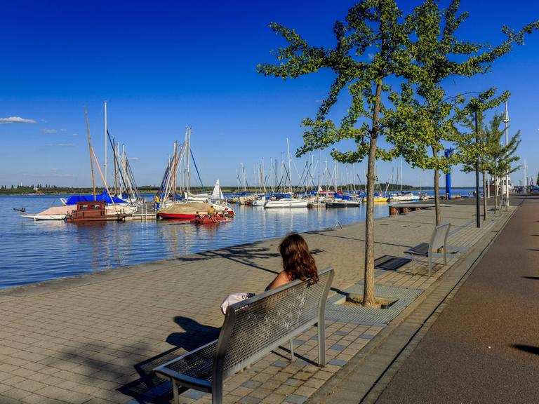 Eine Frau sitzt auf einer Bank an der Marina des Goitzschesees. Im Hintergrund liegen kleine Yachten und Segelboote.