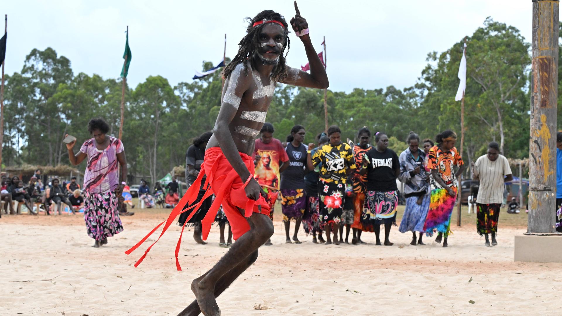 Ein bemalter Mann tanzt in rotem Rock auf einer Sandfläche, im Hintergrund Aboriginee-Frauen.