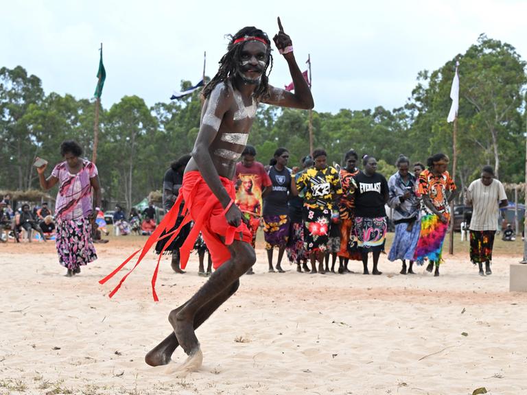 Ein bemalter Mann tanzt in rotem Rock auf einer Sandfläche, im Hintergrund Frauen.