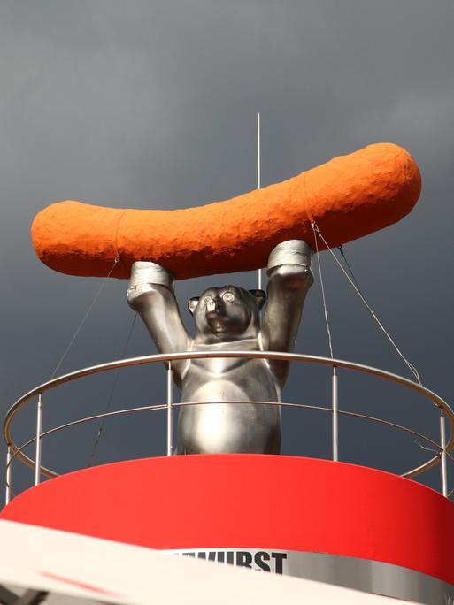 Skulptur eines Bären mit Wurst auf einem Imbiss in Berlin, vor wolkenverhangenem Himmel