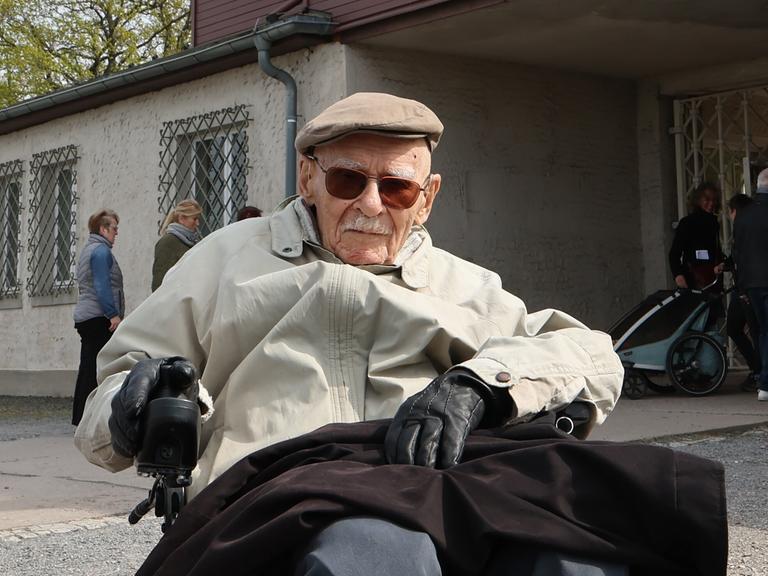 Raymond Renaud aus Frankreich sitzt im Rollstuhl vor einem Tor des ehemaligen NS-Konzentrationslagers Buchenwald.