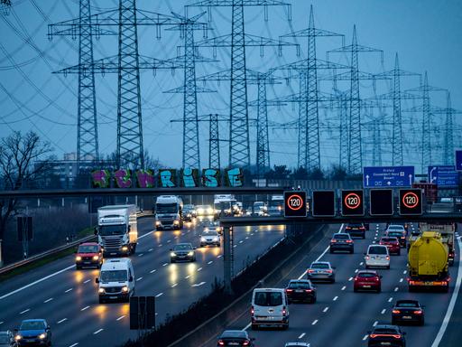 Die dichtbefahrene Autobahn A57 bei Kaarst in der Abenddämmerung. Im Hintergrund stehen viele Stromtrassen vor einem dunklen Himmel.