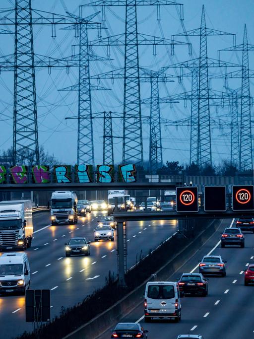 Die dichtbefahrene Autobahn A57 bei Kaarst in der Abenddämmerung. Im Hintergrund stehen viele Stromtrassen vor einem dunklen Himmel.