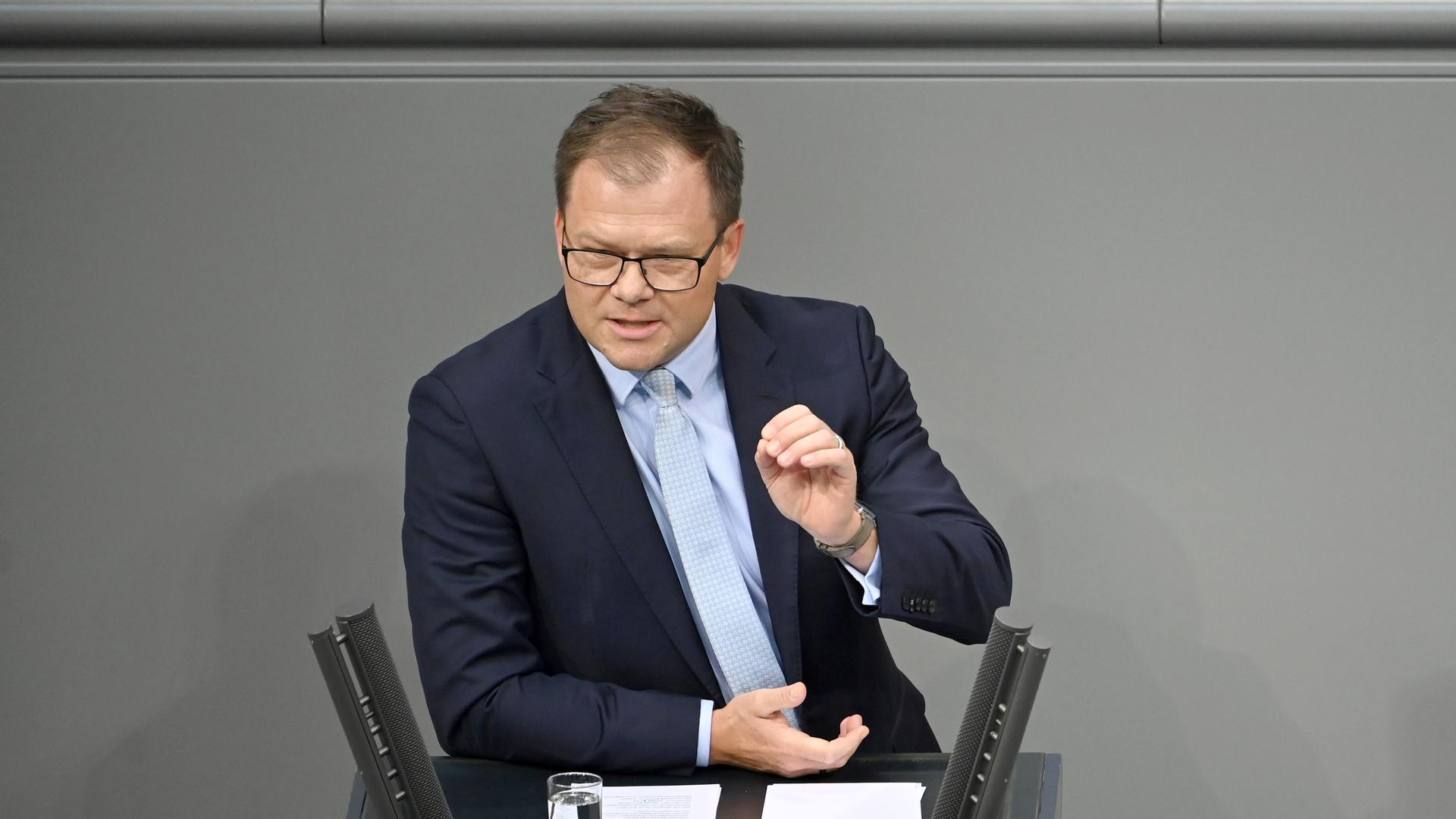 Schneider spricht am Rednerpult zu den Abgeordneten. Er gestikuliert mit der linken Hand. Schneider spricht am Rednerpult zu den Abgeordneten. Er gestikuliert mit der linken Hand.