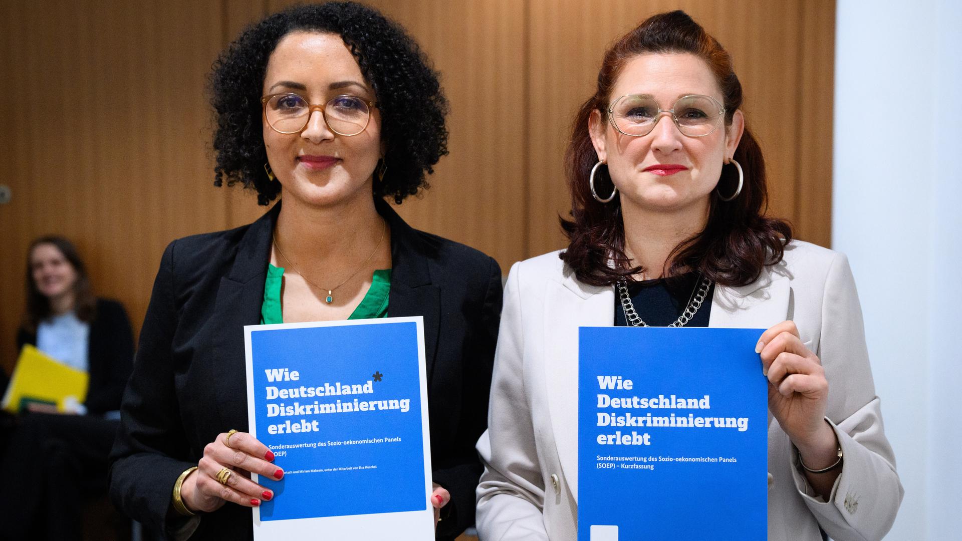 Die beiden stehen lächelnd nebeneinander und halten je eine Version des blauen Berichtshefts vor sich. 