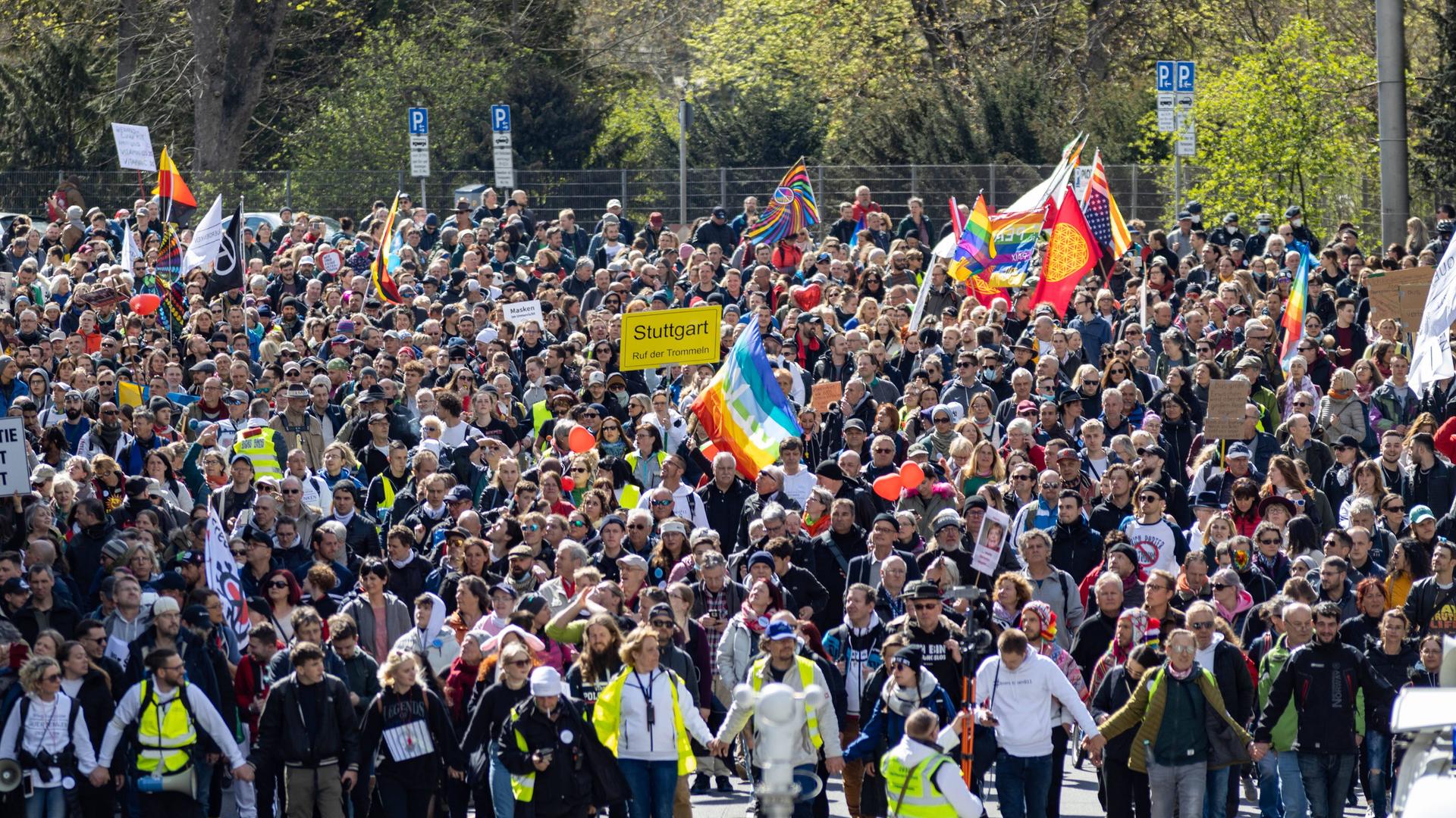Eine Menschenmenge mit Plakaten auf einer Straße in Stuttgart. Menschen in neongelben Westen stehen vorn, einige Demonstranten schwenken Deutschlandfahnen, eine Regenbogenfahne ist zu sehen, andere halten ein Schild im Stil eines Ortschilds hoch, auf dem Stuttgart steht.