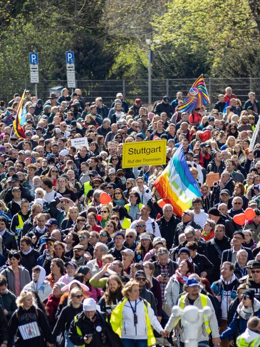 Eine Menschenmenge mit Plakaten auf einer Straße in Stuttgart. Menschen in neongelben Westen stehen vorn, einige Demonstranten schwenken Deutschlandfahnen, eine Regenbogenfahne ist zu sehen, andere halten ein Schild im Stil eines Ortschilds hoch, auf dem Stuttgart steht.