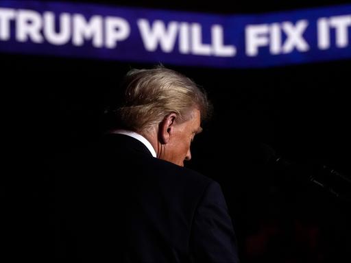 Rückenansicht von Donald Trump auf der Bühne einer Wahlkampfveranstaltung. Im Hintergrund sieht man Schrift auf einem Banner: "Trump will fix it!"