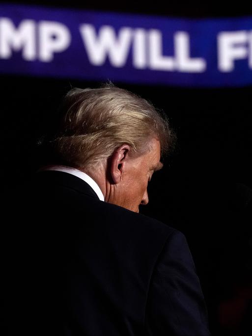 Rückenansicht von Donald Trump auf der Bühne einer Wahlkampfveranstaltung. Im Hintergrund sieht man Schrift auf einem Banner: "Trump will fix it!"