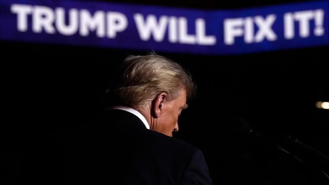 Rückenansicht von Donald Trump auf der Bühne einer Wahlkampfveranstaltung. Im Hintergrund sieht man Schrift auf einem Banner: "Trump will fix it!"