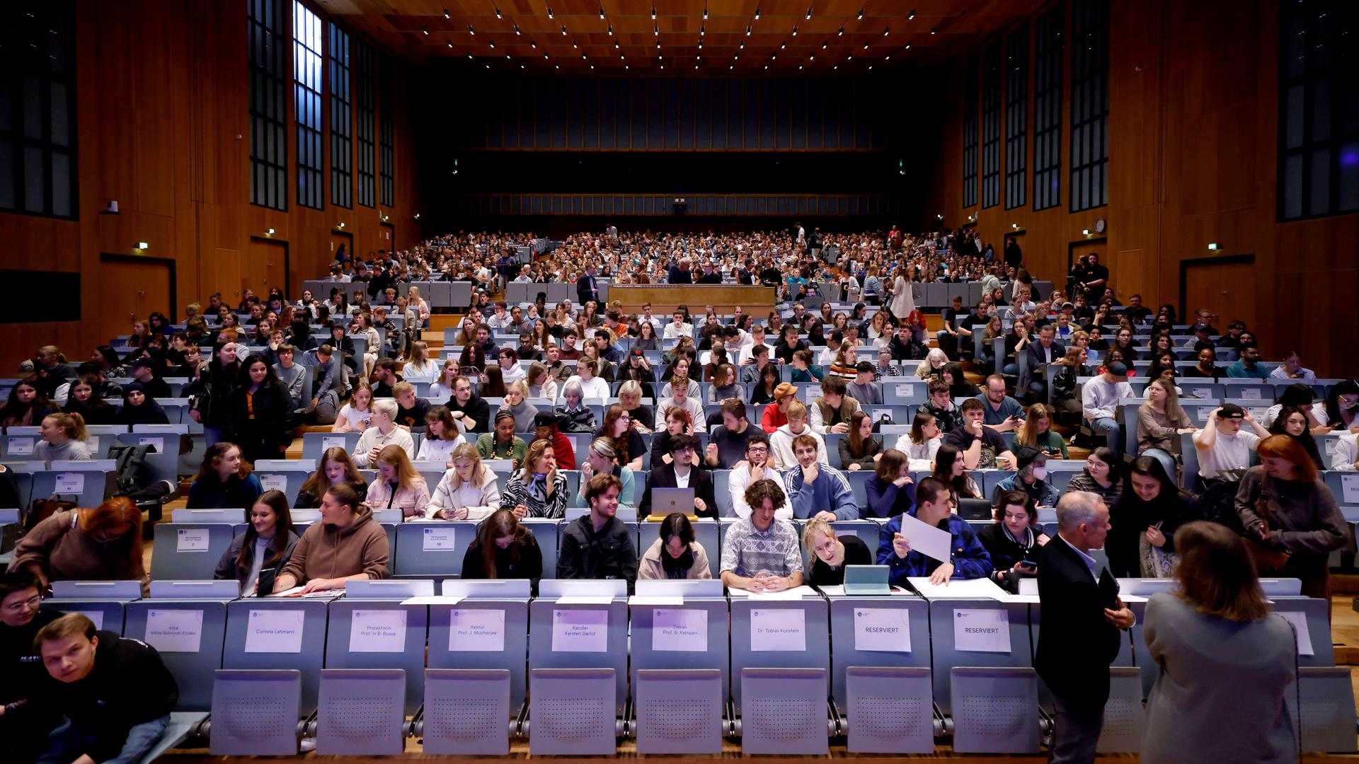 Studierende sitzen in einem großen Hörsaal. In der ersten Reihe sind noch Plätze frei, die mit Namensschildern belegt sind.